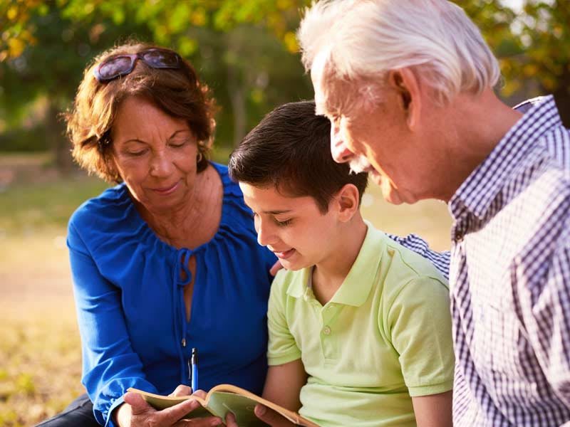 A child reading a book with their grandparents in a park.