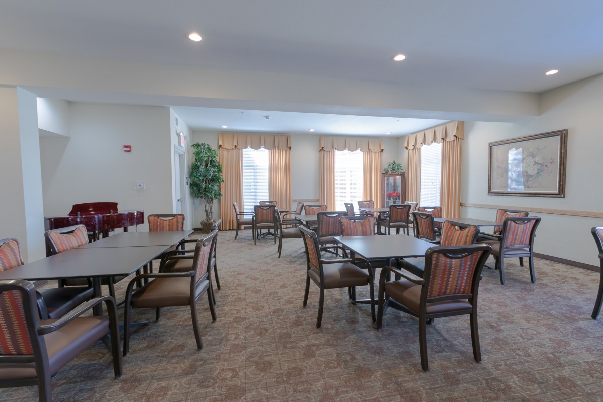 A spacious dining area with multiple tables and chairs, decorated with curtains and carpet, illuminated by overhead lighting.