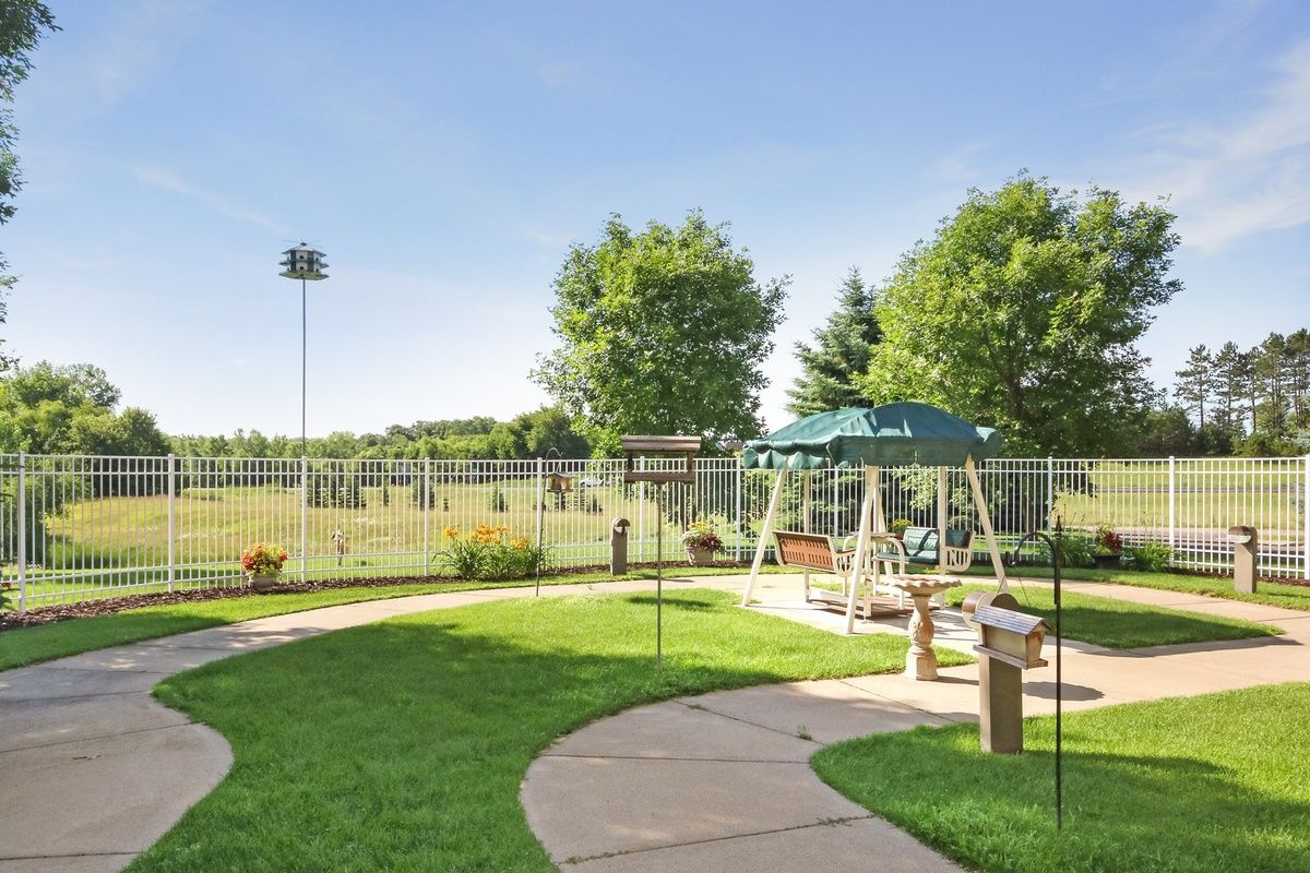 A sunny playground with swings, a pathway, and lush green grass surrounded by trees and a fence.
