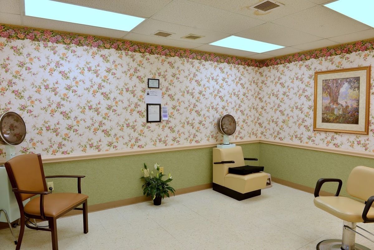 A vintage-style salon waiting area with a floral wallpaper, featuring a hair dryer chair and indoor plant.