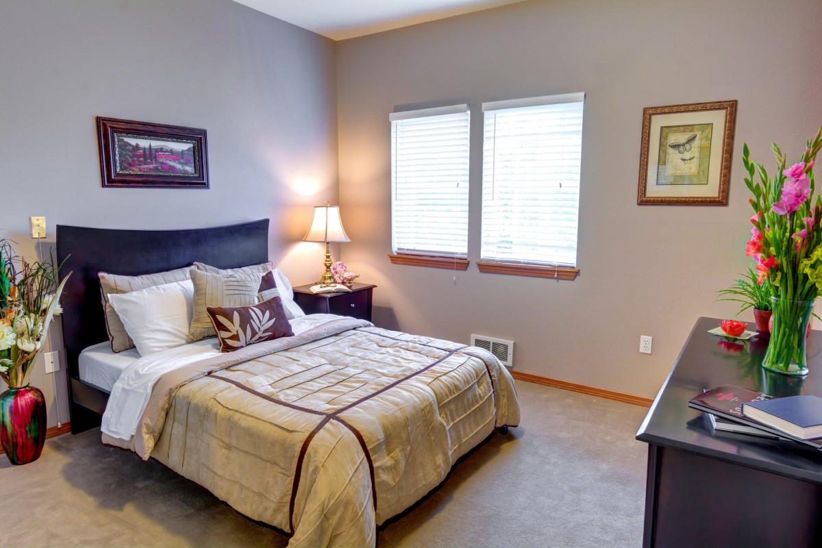 A cozy bedroom with a neatly made bed, bedside table with a lamp, and two windows letting in natural light.