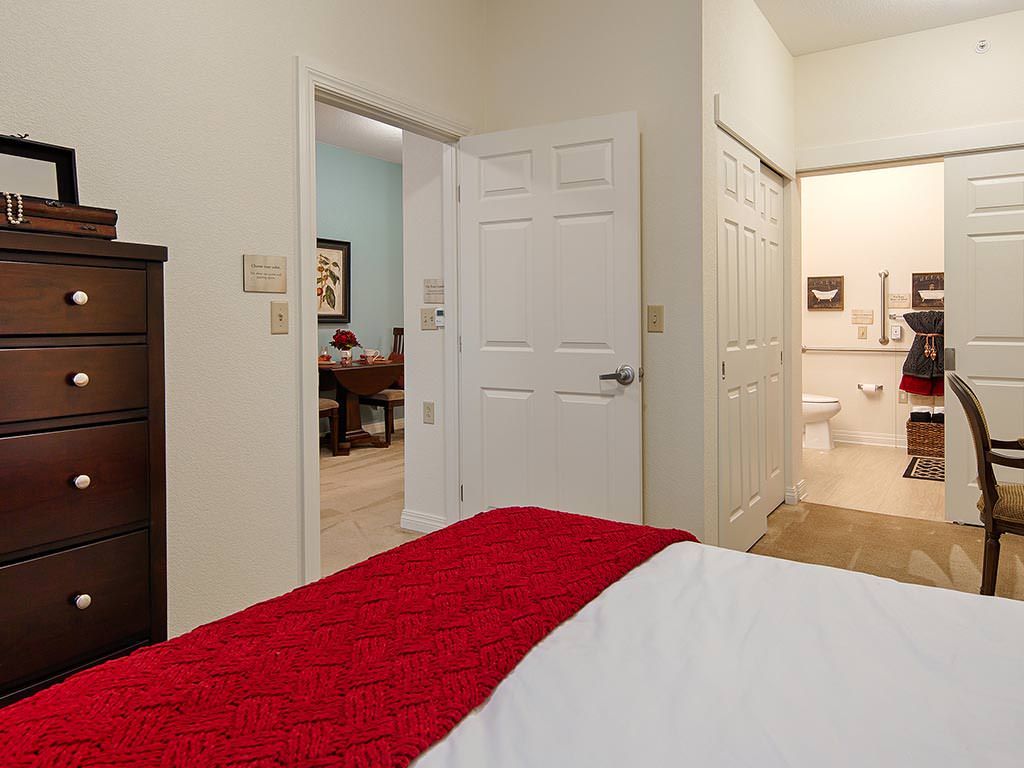 A cozy bedroom with a red blanket on the bed and open doors leading to an office and bathroom.