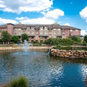 A scenic view of a lakeside hotel with a fountain and lush greenery under a blue sky.