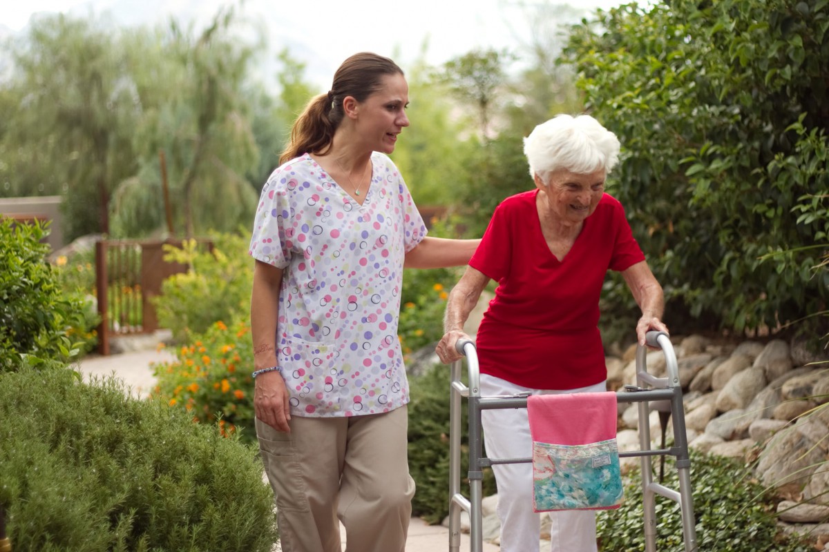 A caregiver assists an elderly woman using a walker during a walk outdoors.