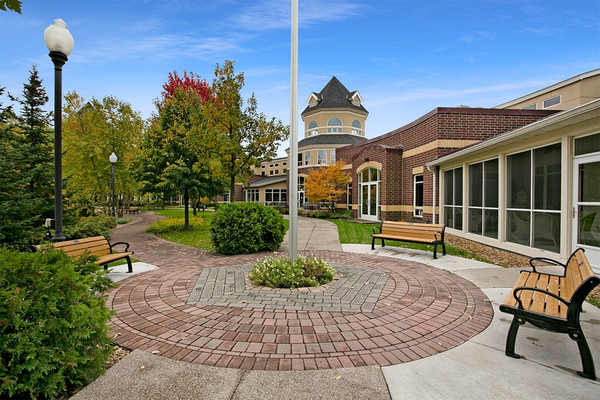 A scenic view of a landscaped garden area with a pathway, surrounded by trees and benches next to a building under a clear blue sky.