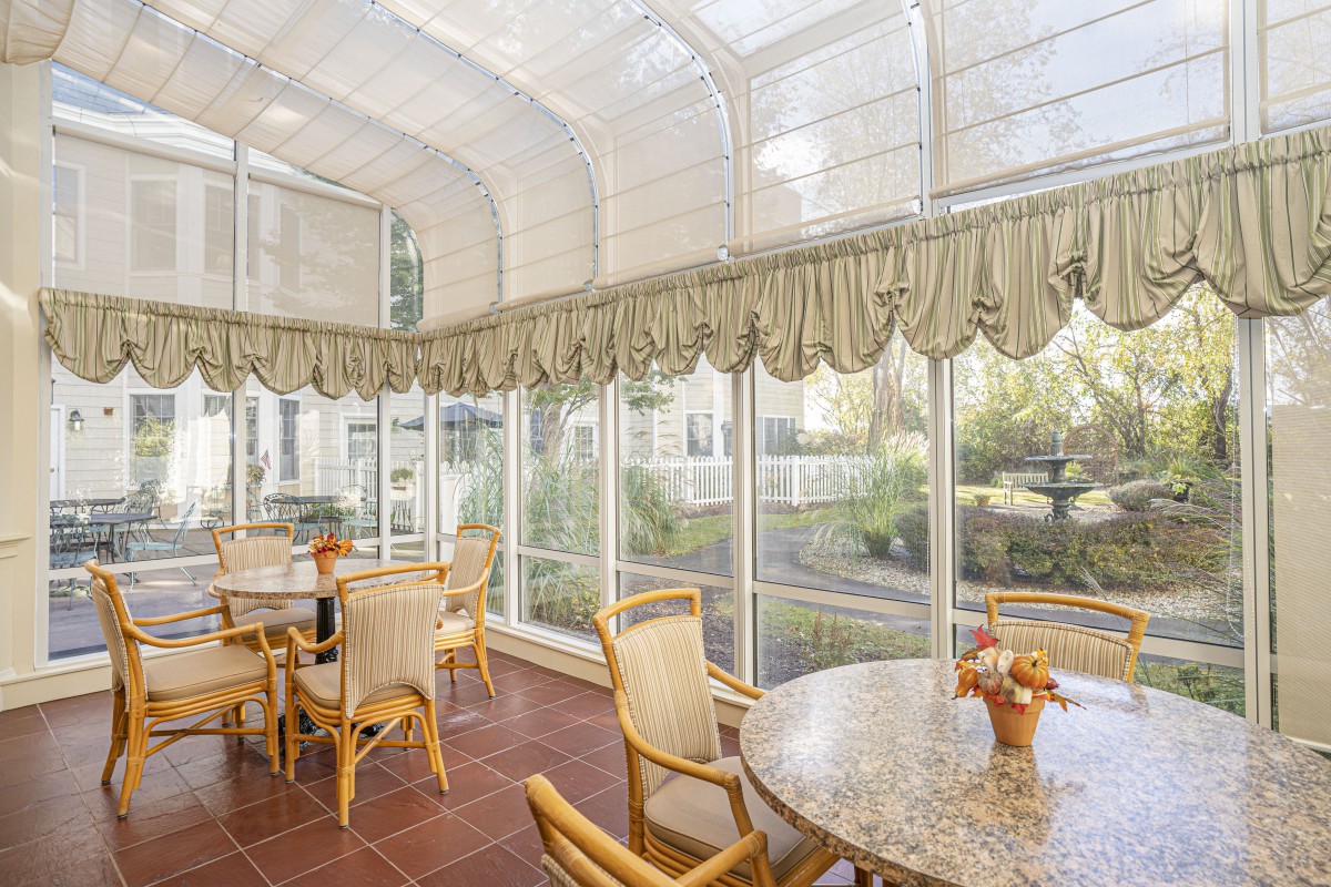 A bright sunroom with glass walls, featuring a dining area with tables and chairs under abundant natural light.