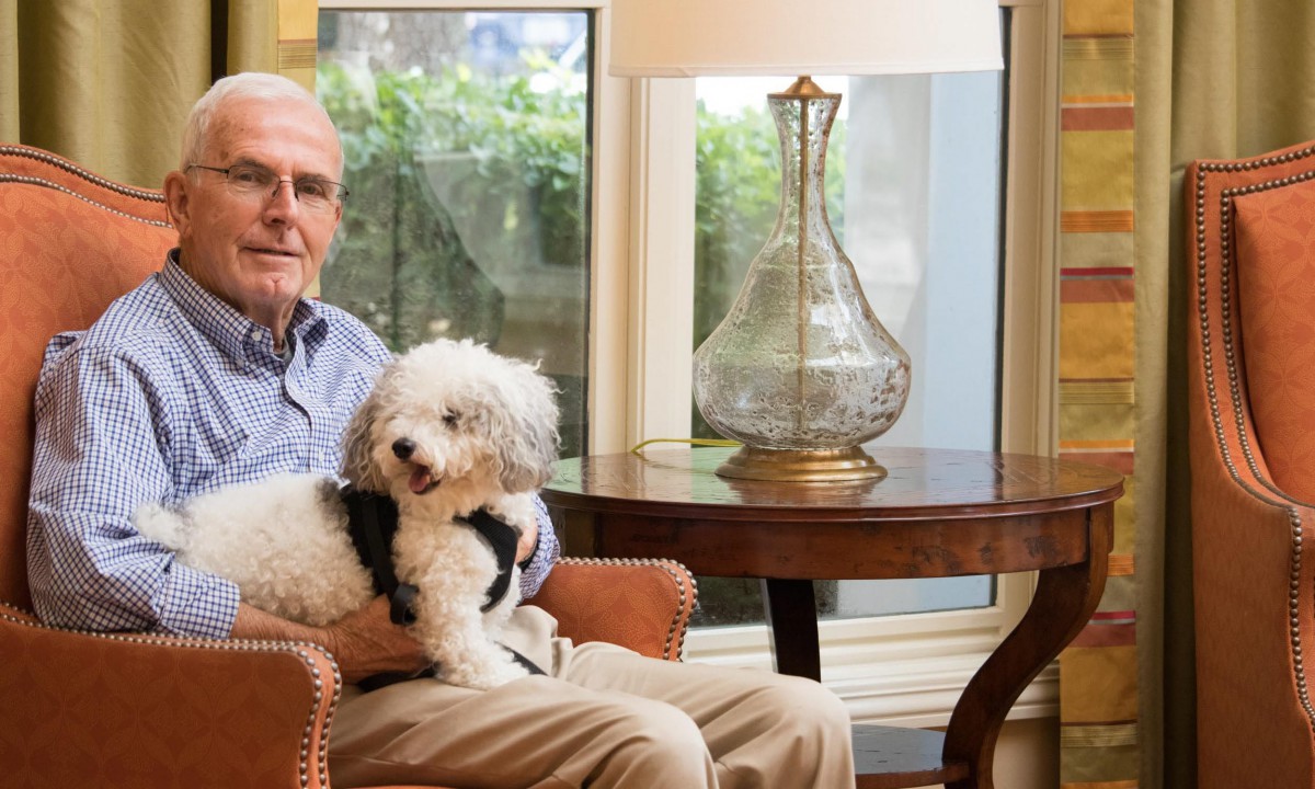 An elderly man sitting in a chair with a small white dog on his lap, next to a lamp and a window.