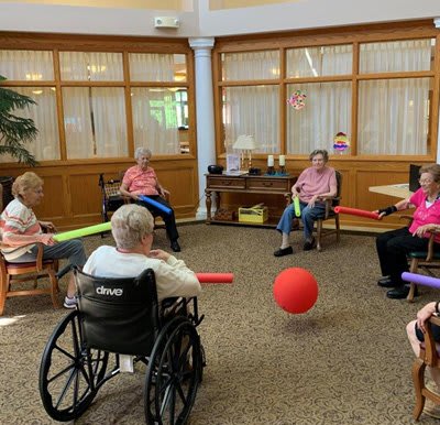 A group of seniors participating in a fun indoor ball game using noodles while seated in a circle.