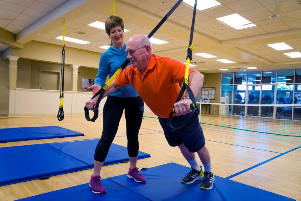 A senior man performs TRX exercises with the guidance of a trainer in a gym setting.