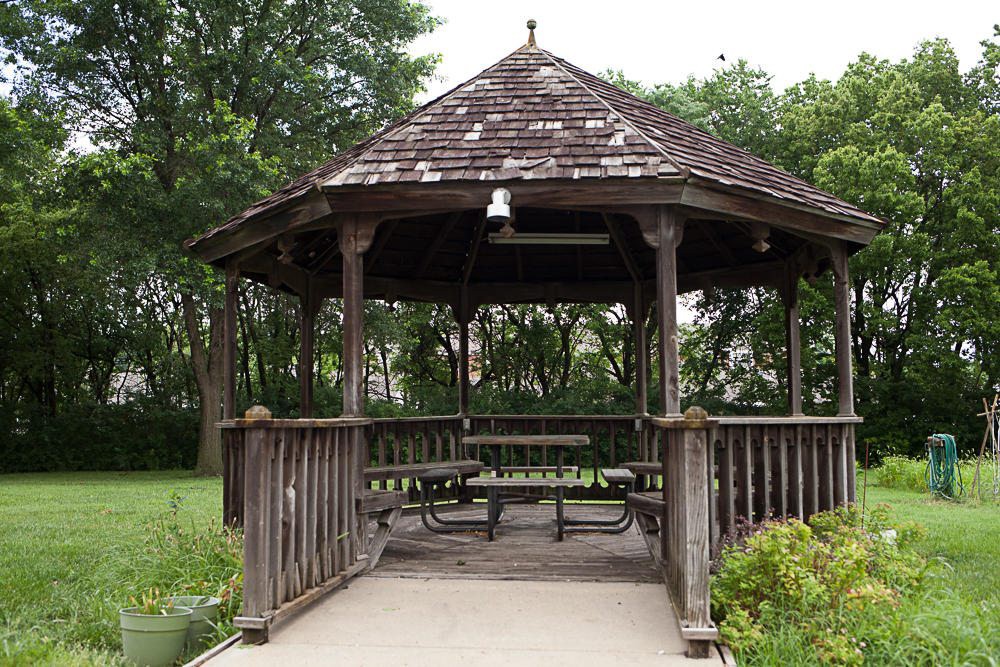 A wooden gazebo with picnic tables, surrounded by greenery in a park setting.