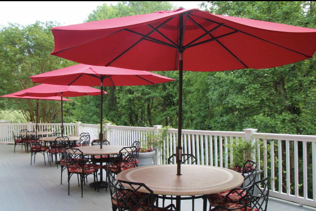 An inviting outdoor patio with red umbrellas and metal tables set against a lush, green backdrop.