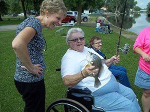 A person in a wheelchair proudly holding a fish they caught, surrounded by others enjoying a day outdoors.