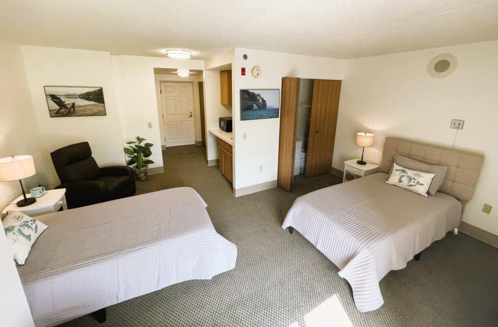 A spacious bedroom featuring two twin beds with neutral-toned bedding, a lamp on a small table, and simple furniture in a carpeted setting.