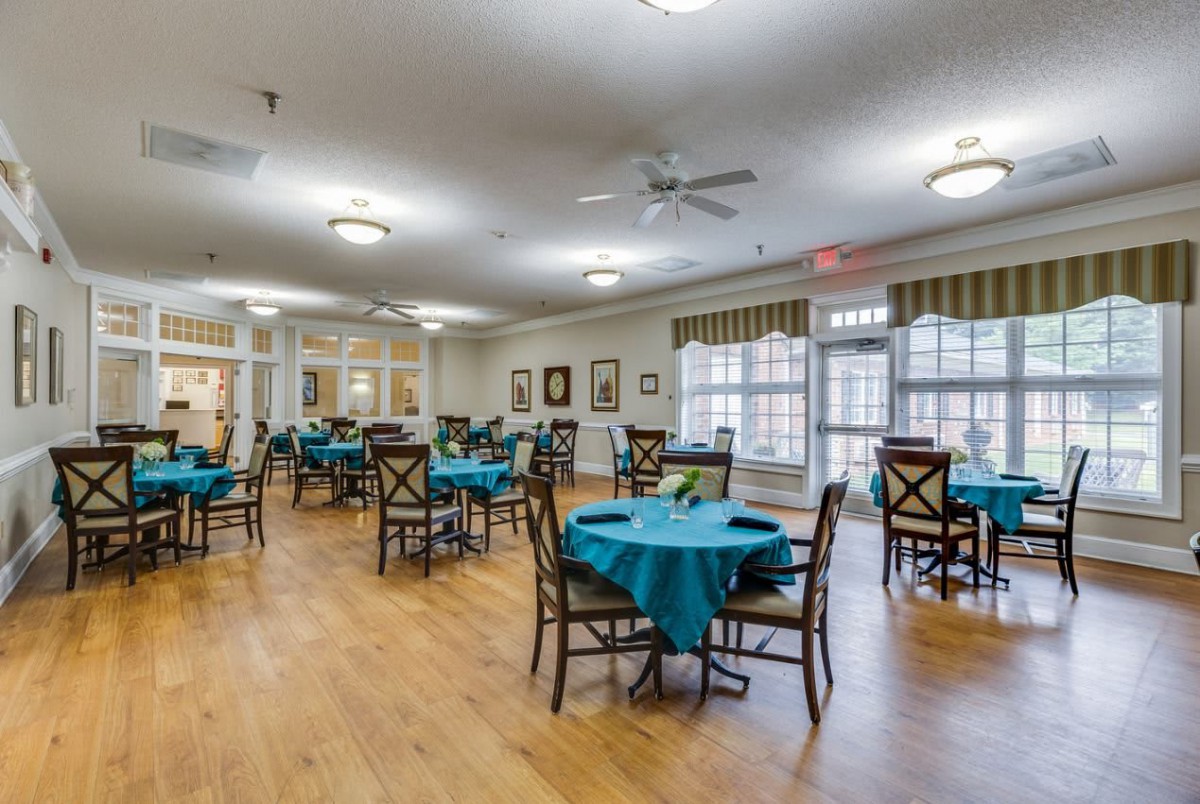 A spacious dining room with multiple tables and chairs, featuring teal tablecloths and large windows allowing natural light.