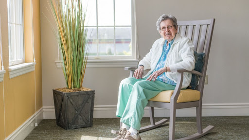 An elderly woman smiles while sitting comfortably in a rocking chair, surrounded by natural light and greenery.