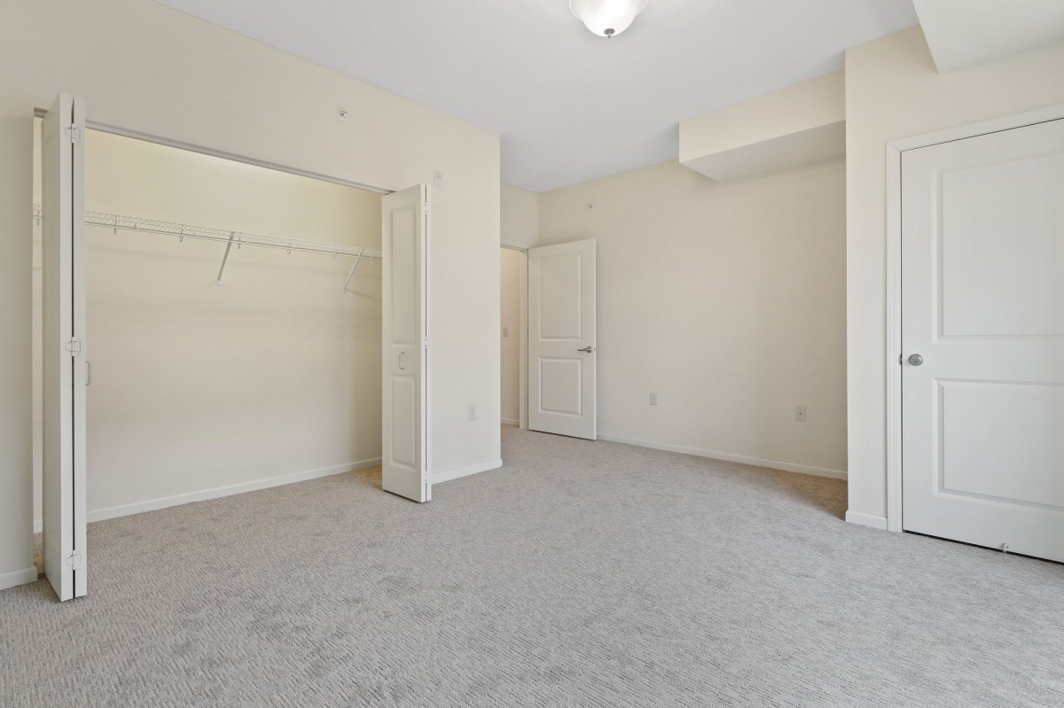 An empty room with neutral walls and carpet, featuring an open closet and a closed door.