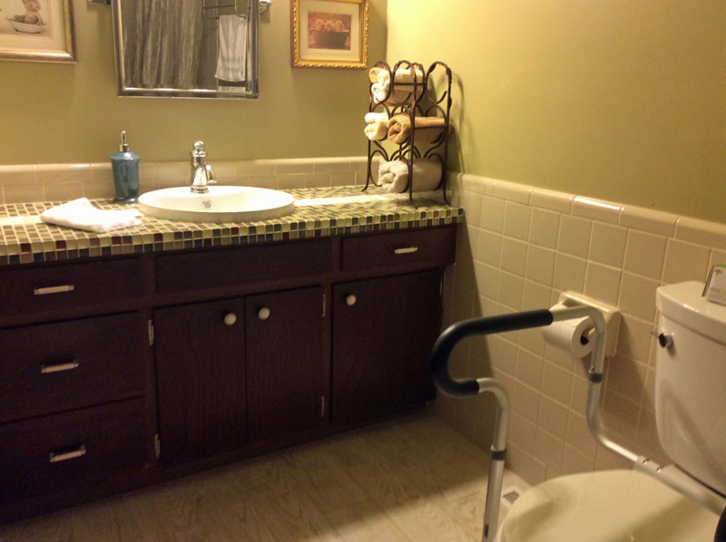 A bathroom featuring a sink and countertop with a decorative towel rack, mirror, and handicap accessible toilet seat.