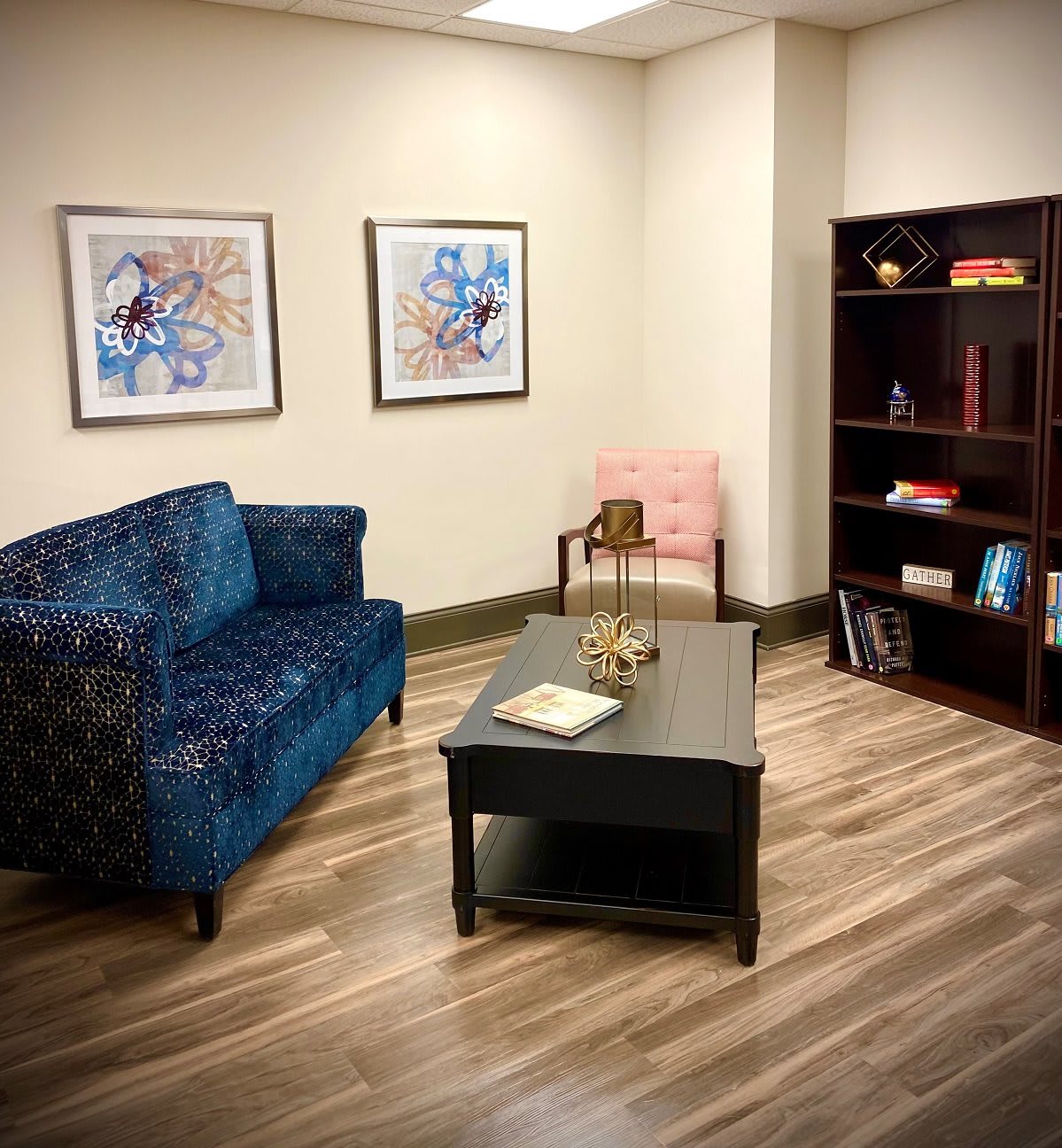 A cozy office with a blue sofa, two framed artworks, and a bookshelf on a wooden floor.