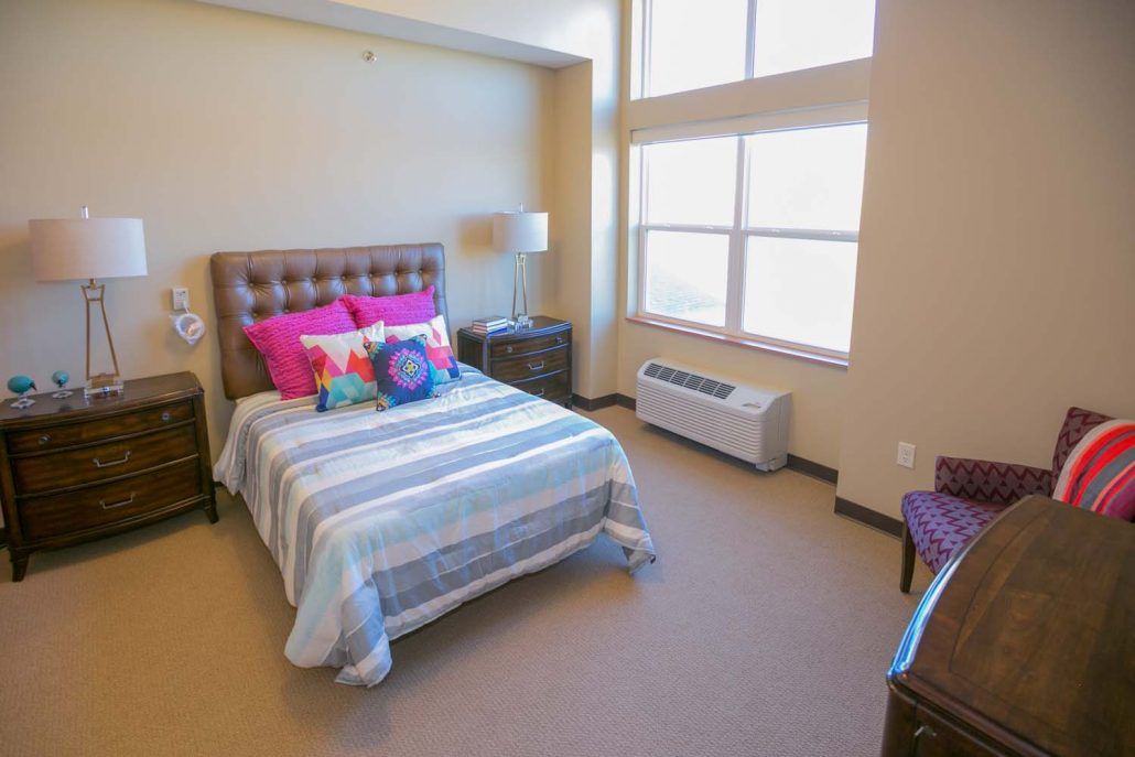 A cozy bedroom with a bed, colorful pillows, and large windows.