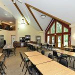 A spacious dining hall with multiple tables and chairs, featuring a high ceiling and large windows.