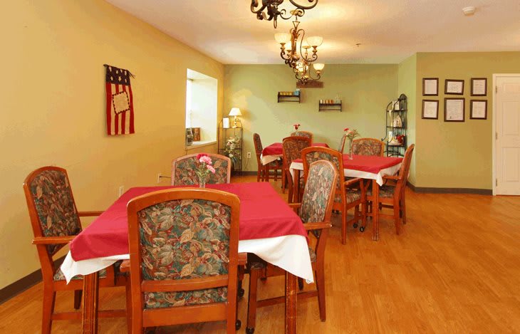 A cozy dining room with multiple tables set with red tablecloths and patterned chairs, illuminated by a chandelier.