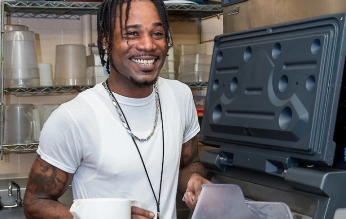 A smiling person holding a cup, standing indoors near equipment.