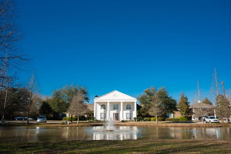 A large white house with columns stands behind a pond and fountain, surrounded by trees under a clear blue sky.