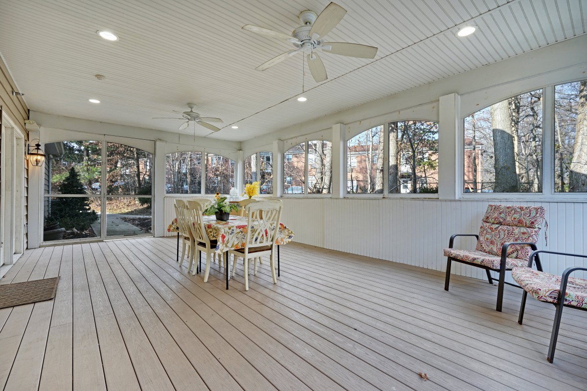 A spacious porch with a dining area, featuring ceiling fans and wood flooring, surrounded by windows.