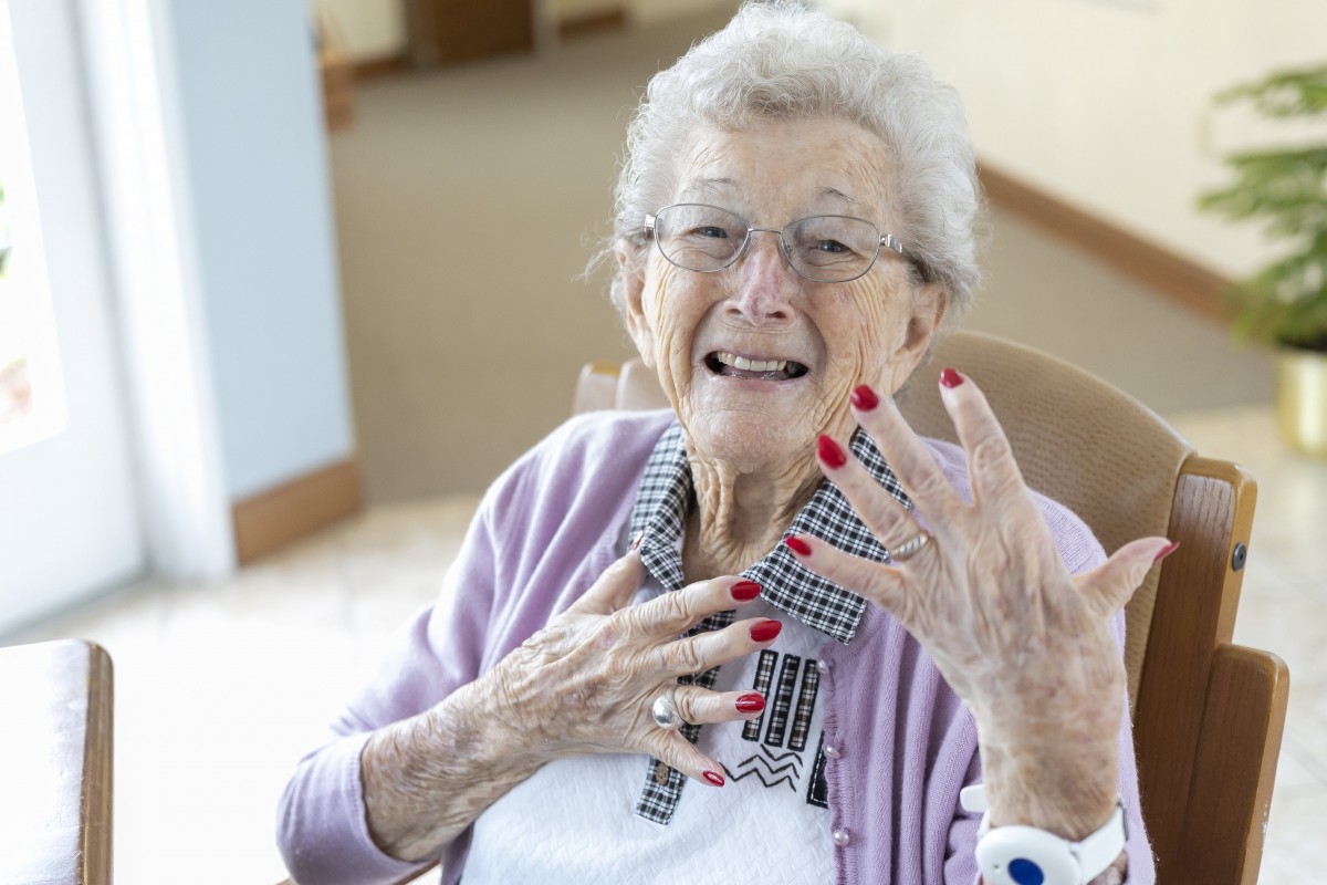 An elderly woman with glasses smiling and showing off her red nails indoors.