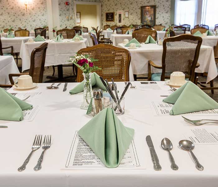 An elegantly set dining room with neatly arranged tables featuring green napkins and polished utensils.