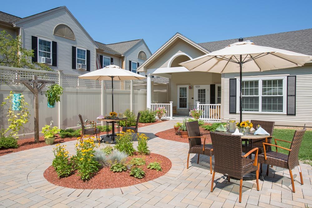 A sunny patio with tables and chairs under umbrellas, surrounded by a beautifully landscaped garden.
