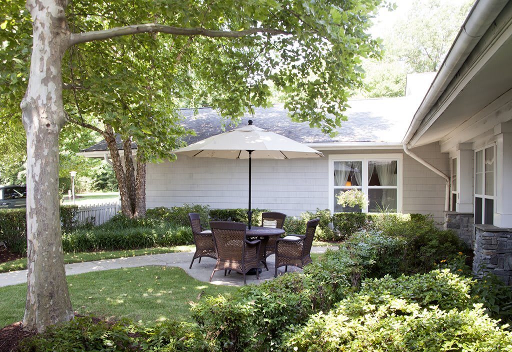 A serene backyard patio featuring outdoor seating under a large umbrella, surrounded by lush greenery and well-maintained landscaping.