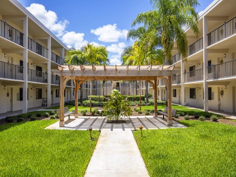 A beautifully landscaped apartment complex courtyard featuring a pergola and palm trees on a sunny day.