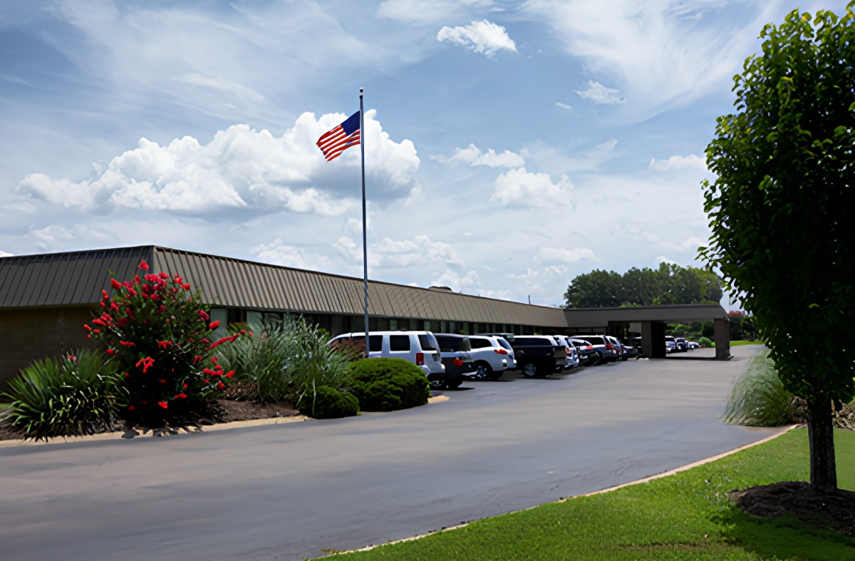 A building with a parking lot and an American flag flying in the foreground, surrounded by landscaped greenery under a partly cloudy sky.