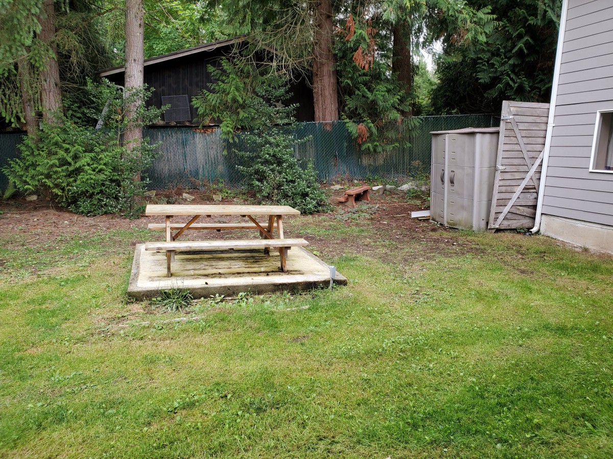 A backyard scene with a wooden picnic table, a grassy area, a small shed, and trees in the background.