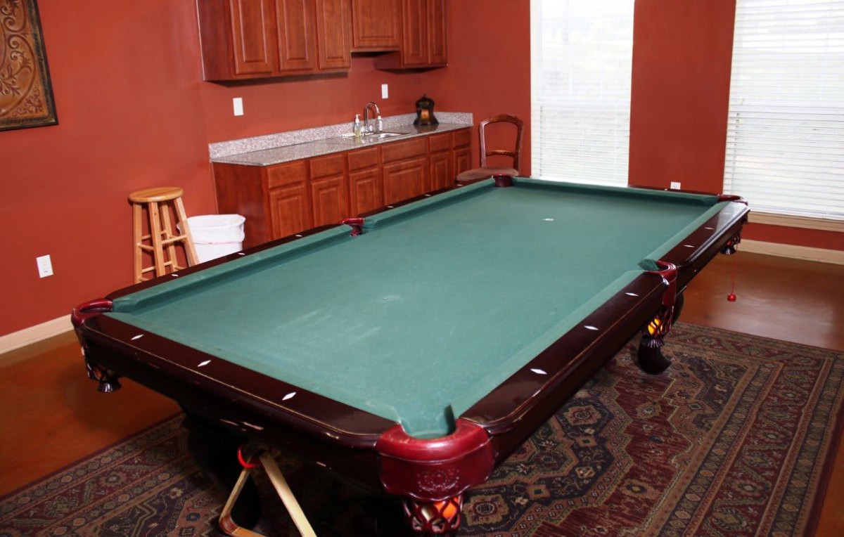 A game room featuring a pool table with a rich wooden finish and green felt, complemented by cabinets in the background.