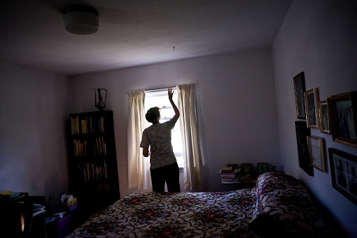 A person standing in a bedroom, drawing back the curtains to let in light.