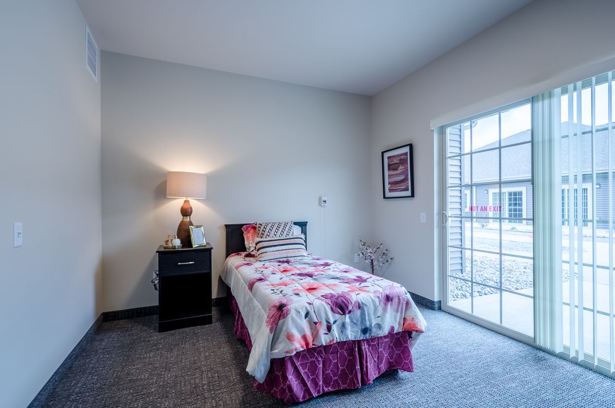 A cozy bedroom featuring a single bed with floral bedding, a nightstand with a lamp, and a large window.