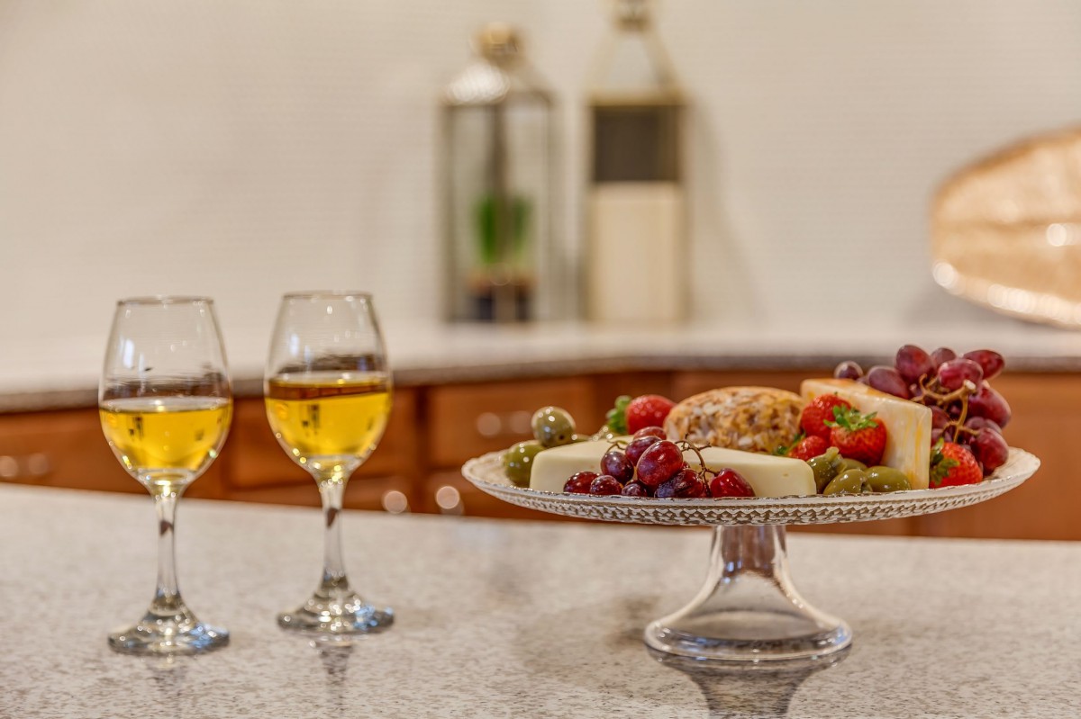 A gourmet platter with cheese and fruits paired with two glasses of white wine on a kitchen counter.