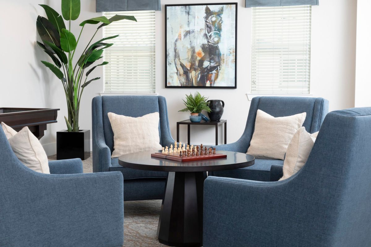 A modern living room with blue chairs centered around a chessboard on a round table, complemented by artwork and an indoor plant.