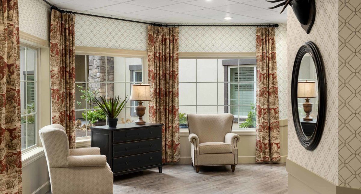 A cozy living room with armchairs, large windows, floral curtains, and a lamp on a dresser.