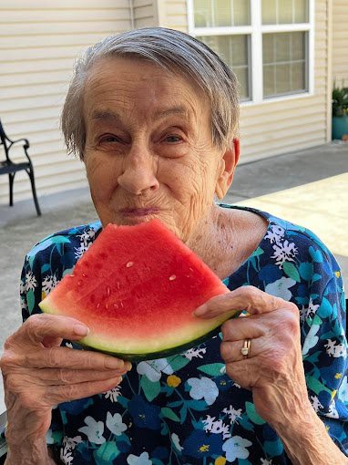 An elderly woman enjoys a slice of watermelon outside on a sunny day.