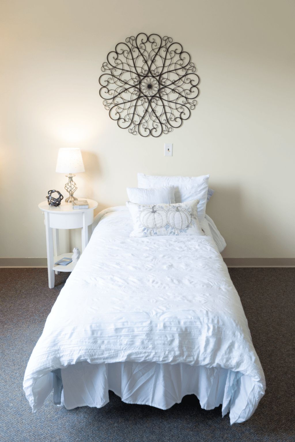 A minimalist bedroom featuring a single bed with white bedding, a nightstand with a lamp, and decorative wall art.