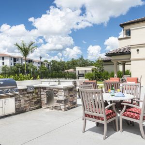 A sunny backyard patio featuring a barbecue grill, outdoor table, and chairs.