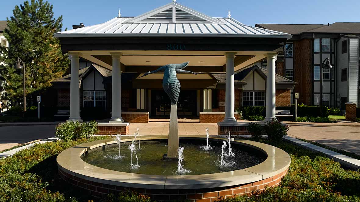 An elegant fountain with a sculpture stands in front of a building with a covered entrance and columns.