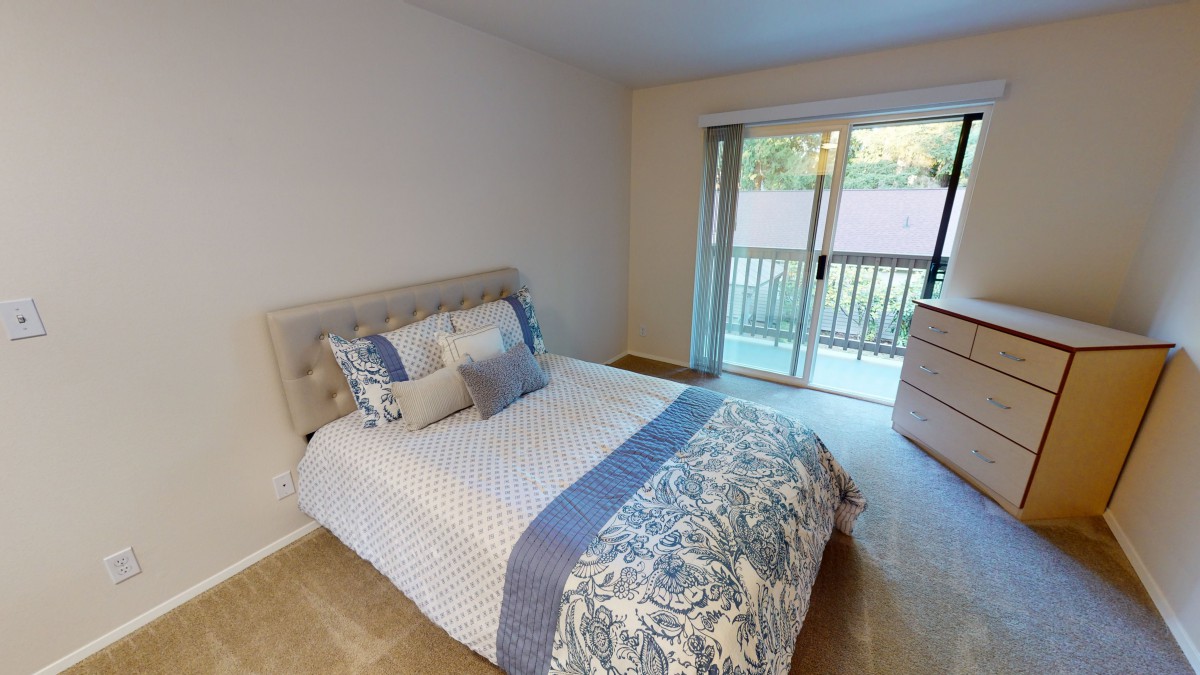 A cozy bedroom featuring a neatly made bed with patterned bedding and pillows, a sliding door leading to a balcony, and soft carpeting.