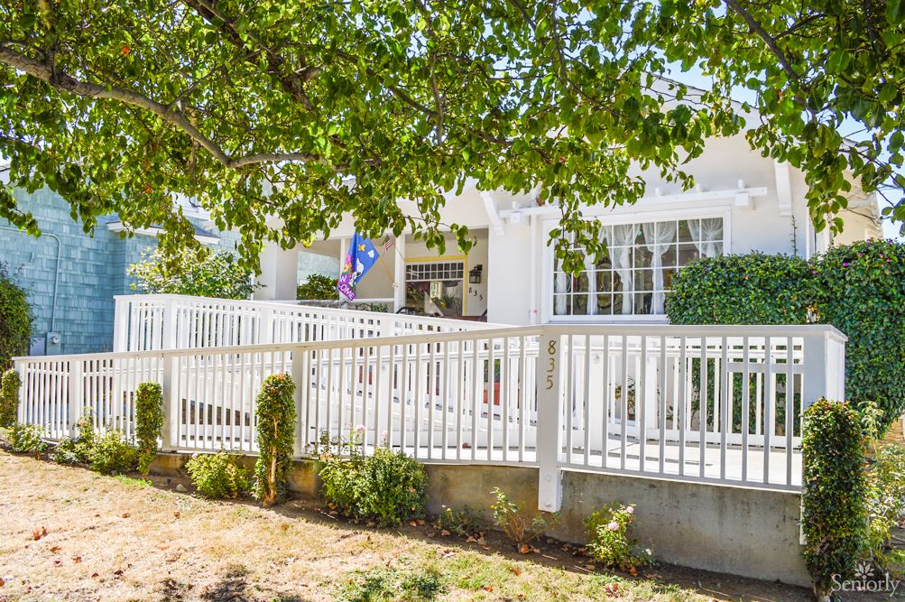 A charming white residential house surrounded by trees, featuring a porch and white fence.