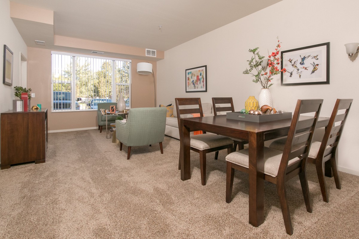 A cozy living and dining area with carpeted flooring, featuring a wooden dining table and chairs, cushioned seating, and large windows allowing natural light.