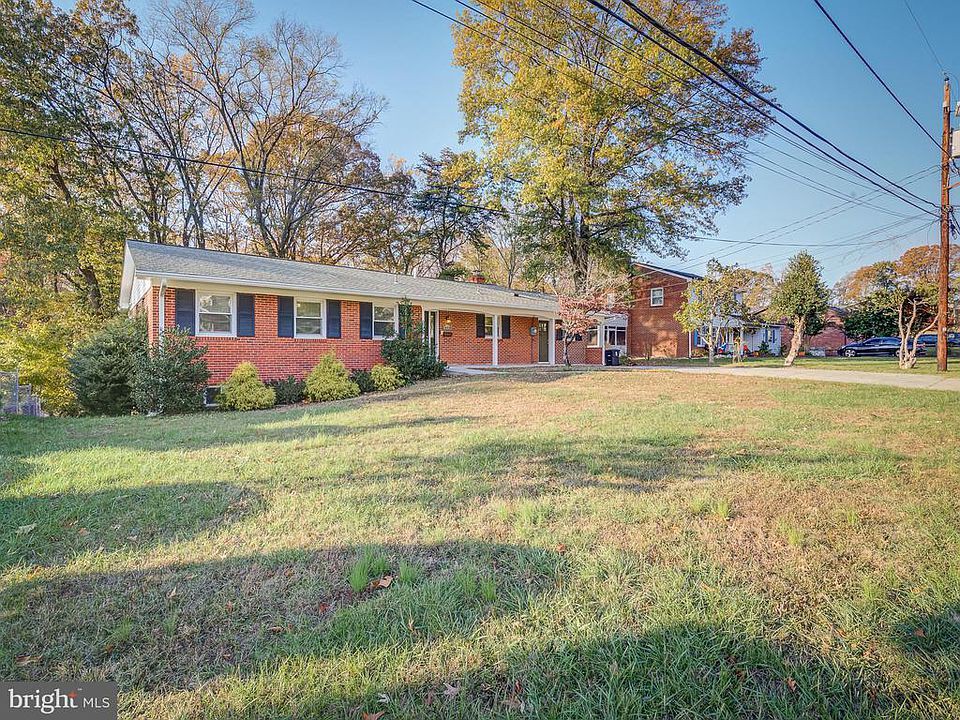 A brick house in a suburban neighborhood with a large yard and trees under a clear, sunny sky.