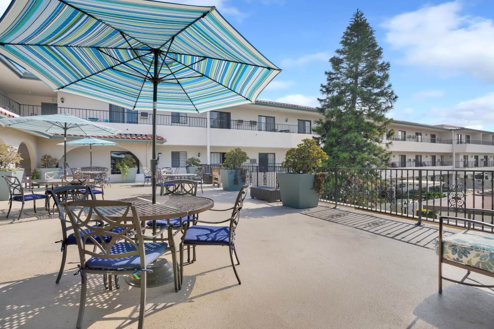 A spacious outdoor patio area with tables, chairs, and umbrellas located outside a multi-story building on a clear day.